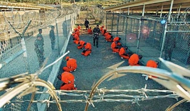 In this photo released 18 January 2002 by the Department of Defense, Al-Qaeda and Taliban detainees in orange jumpsuits sit in a holding area under the surveillence of US military police at Camp X-Ray at Naval Base Guantanamo Bay, Cuba, during in-processi