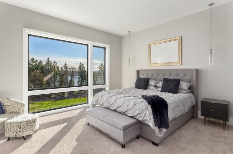 Bedroom in new modern luxury home with very large window inviting natural light. Shows exterior view of blue sky, grass, trees, and water. Bedroom in new modern luxury home with very large window inviting natural light. Shows exterior view of blue sky, grass, trees, and water.